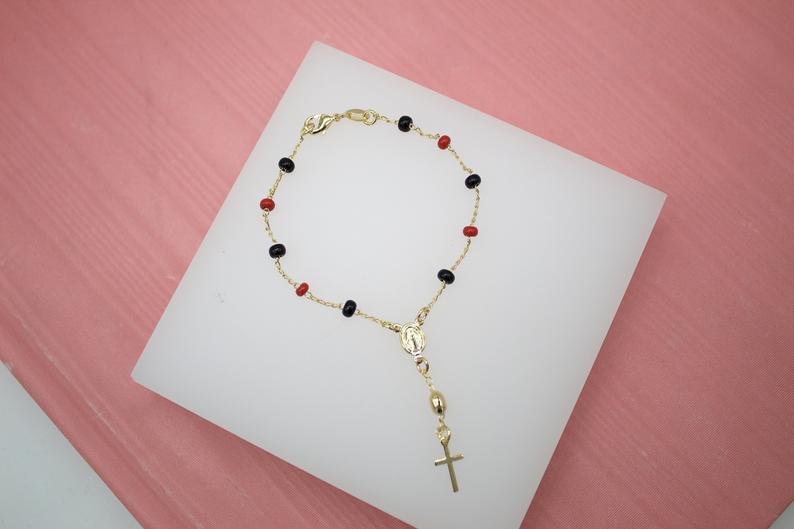 A delicate gold rosary bracelet features alternating black and red beads, a small Miraculous Medal, and a hanging cross charm displayed on a white platform against a pink background.