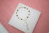 A delicate gold rosary bracelet features alternating black and red beads, a small Miraculous Medal, and a hanging cross charm displayed on a white platform against a pink background.