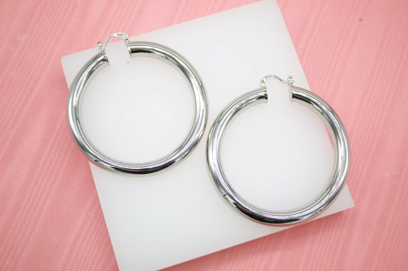 A pair of polished silver hoop earrings rests on a white display block against a textured pink background.