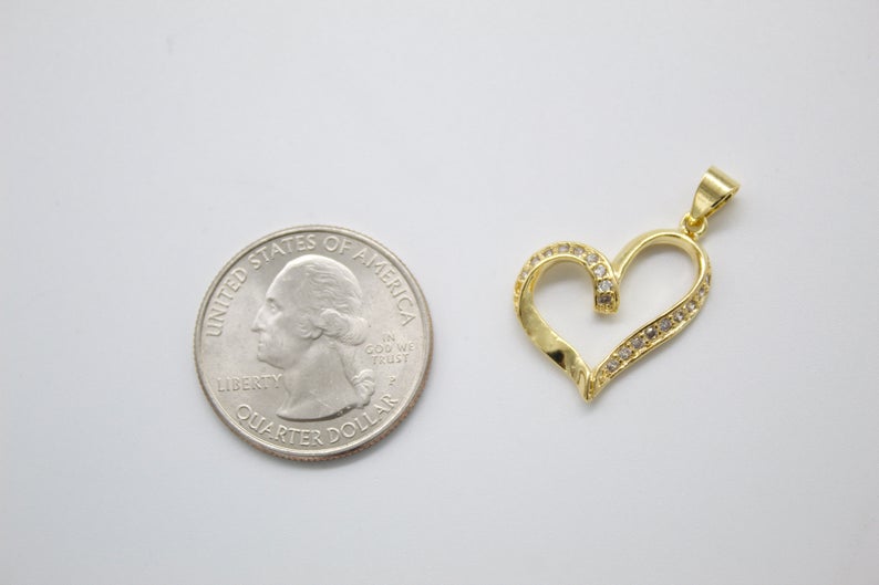 A gold heart-shaped pendant studded with tiny diamonds rests beside a US quarter coin on a white surface to show its size.