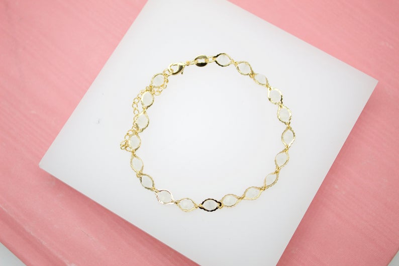 A delicate gold link bracelet rests on a white display surface against a pink background.