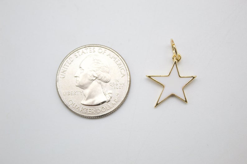 A gold-plated star charm with white enamel rests next to a US quarter coin for scale on a light gray surface.