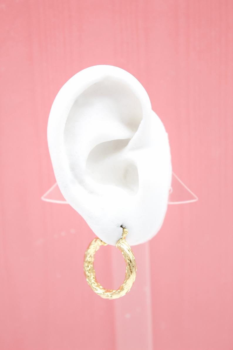 A textured gold hoop earring hangs from a white mannequin ear against a pink background.
