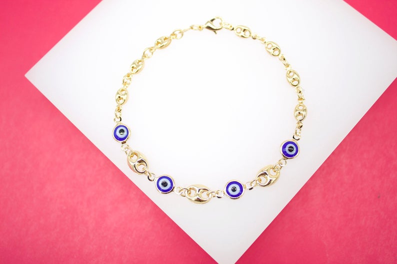 A gold chain bracelet featuring blue evil eye charms rests on a white display against a vibrant pink background.
