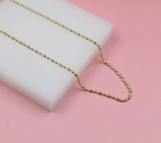 A delicate gold bead chain necklace rests on a white display block against a pink background.