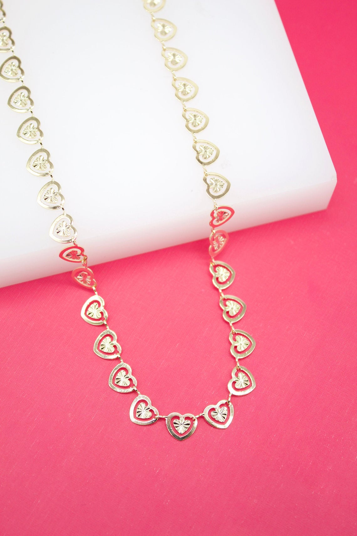 A delicate gold heart-link chain necklace rests elegantly on a white display block against a vibrant pink background.
