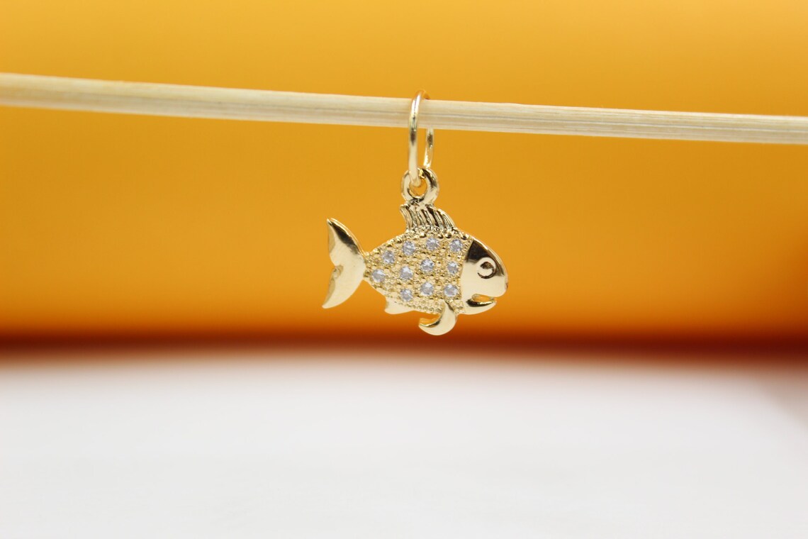 A gold fish pendant with sparkling cubic zirconia stones hangs delicately from a wooden stick against a bright orange and white backdrop.