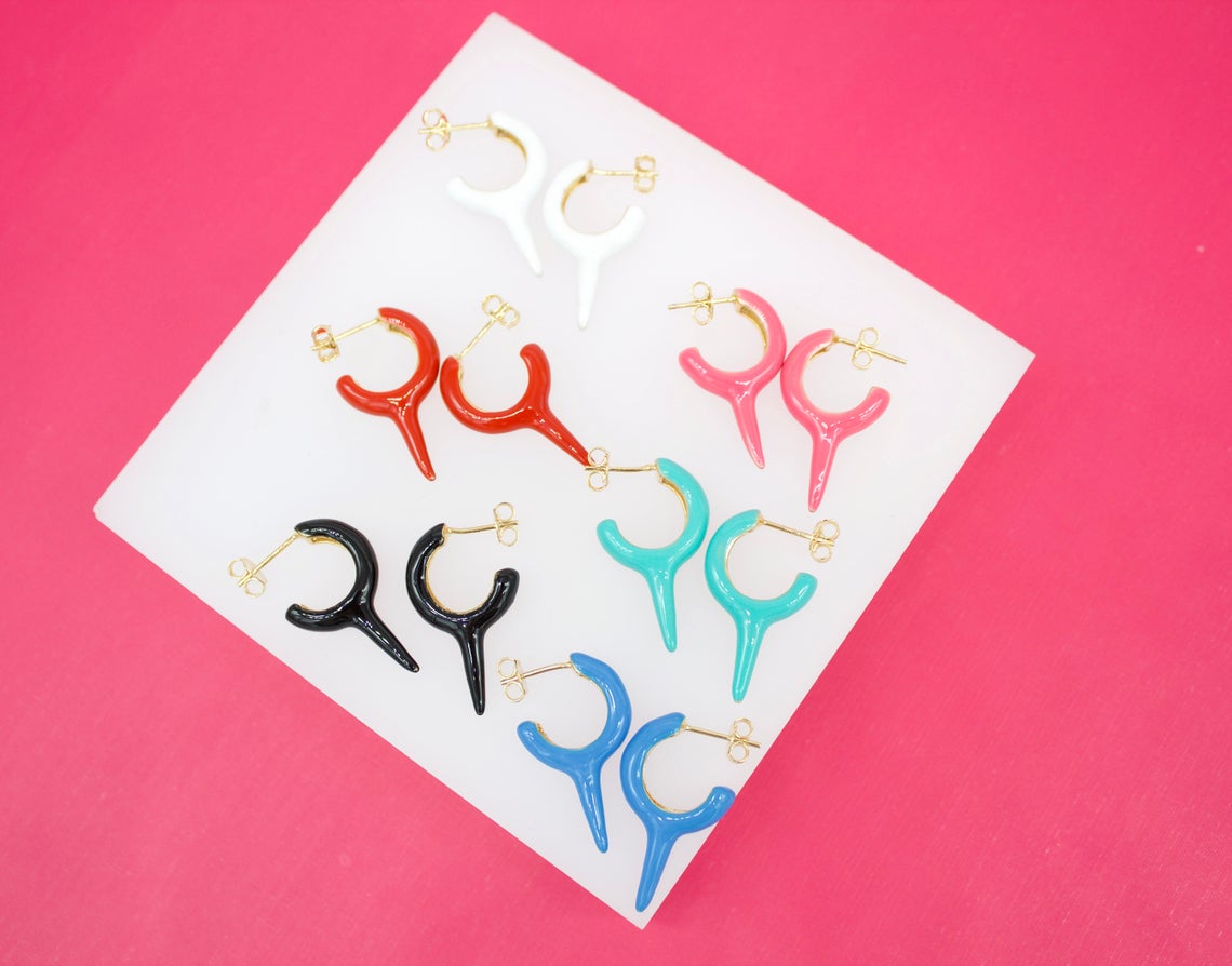 An assortment of colorful horn-shaped enamel earrings in white, red, pink, black, turquoise, and blue is displayed on a white card against a vibrant pink background.