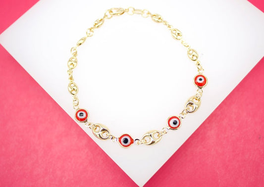 A gold chain bracelet features delicate red and black evil eye charms displayed on a white and pink background.