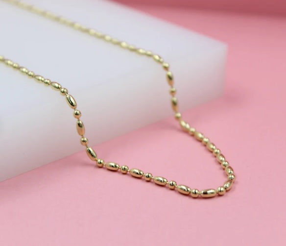 A delicate gold bead and oval-link chain necklace rests elegantly on a soft pink and white display surface.