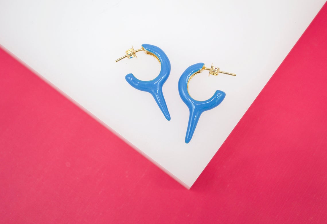 A pair of vibrant blue enamel hoop earrings with a unique spike design rests on a white and pink surface.