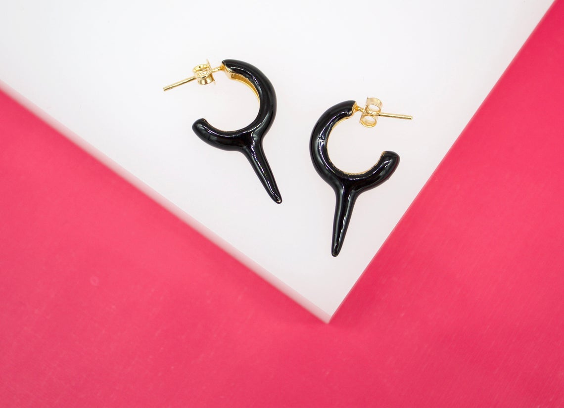A pair of glossy black huggie hoop earrings featuring a distinct downward-pointing thorn charm rests on a white and pink display surface.