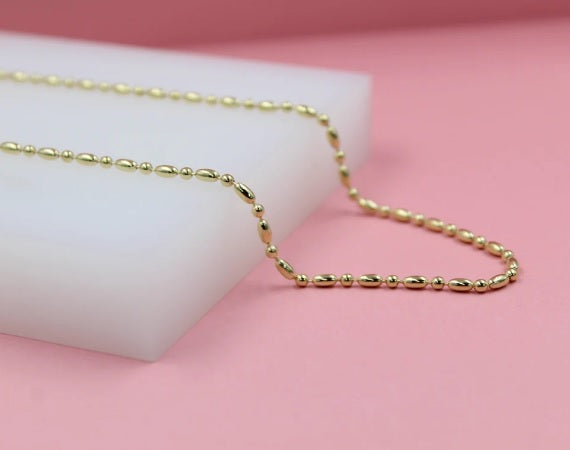 A delicate gold chain necklace with alternating round and oval beads rests on a white display block against a soft pink background.