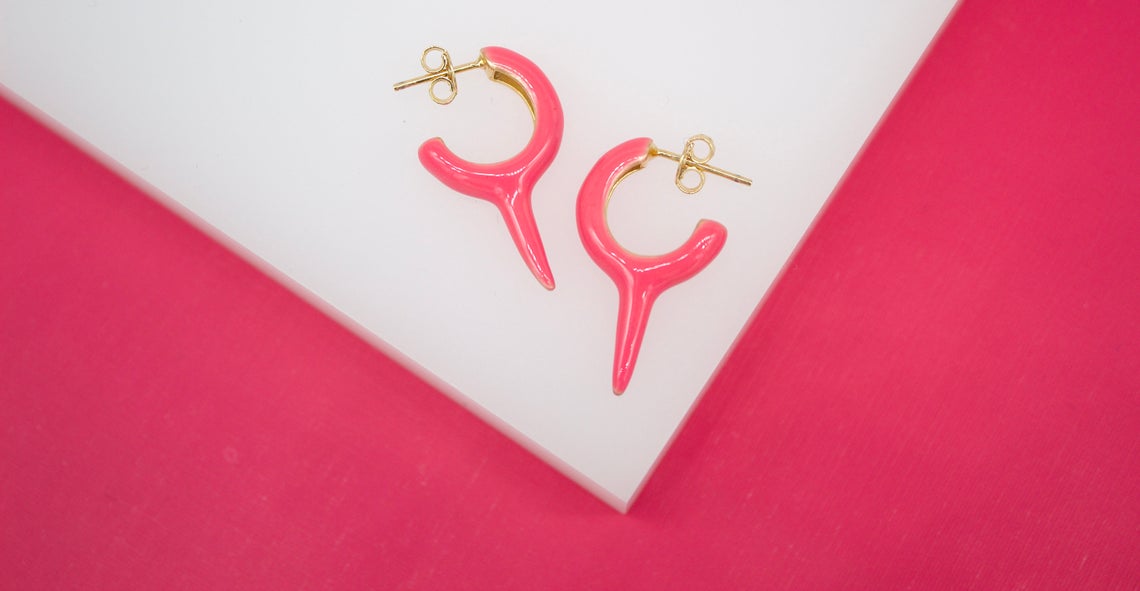 A pair of vibrant pink enamel hoop earrings with gold-plated details rests on a white surface against a bright pink background.