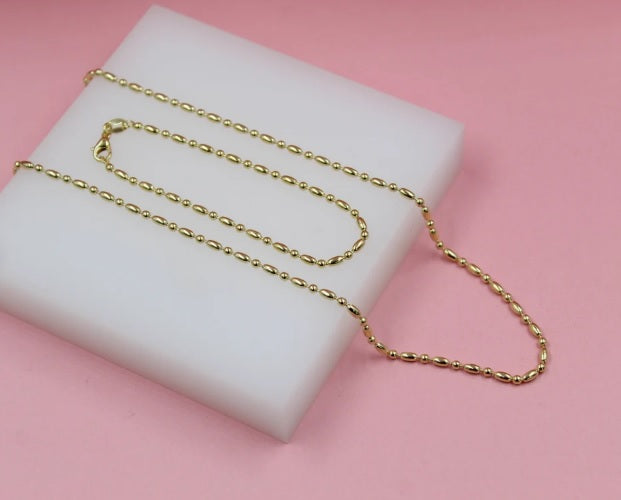 A delicate gold bead chain necklace rests on a white display block against a soft pink background.