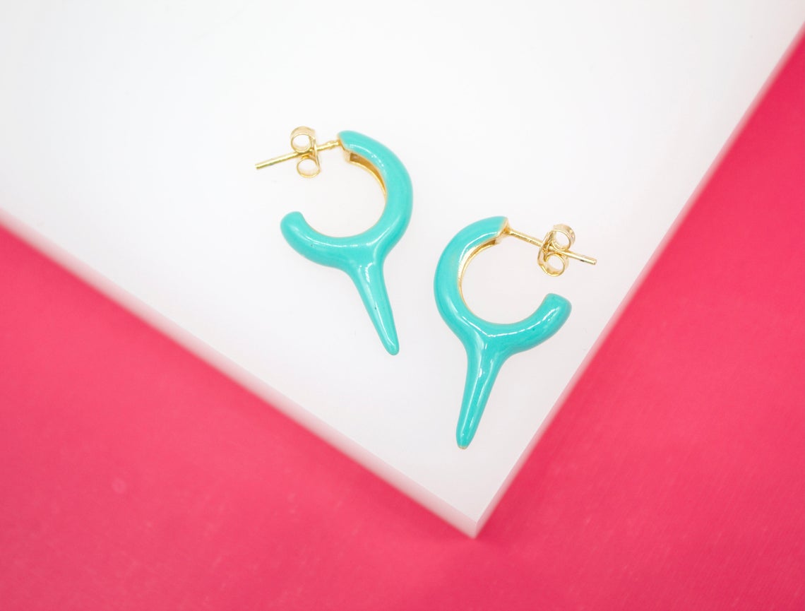 A pair of turquoise enameled huggie hoop earrings with spike charms rest on a white surface against a bright pink background.