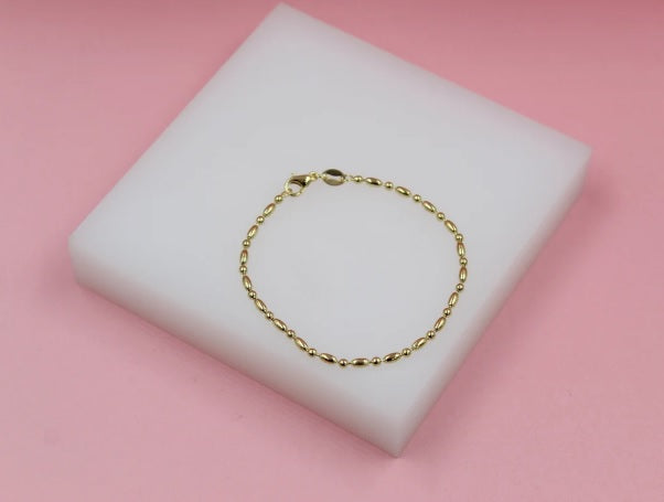 A delicate gold bead bracelet rests on a white display block against a soft pink background.