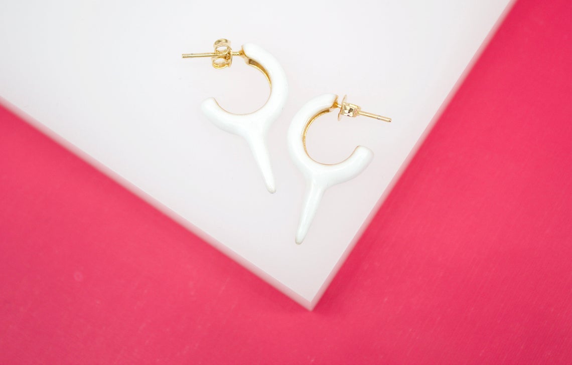 A pair of white enamel hoop earrings with gold-plated posts rests on a white display block against a vibrant pink background.
