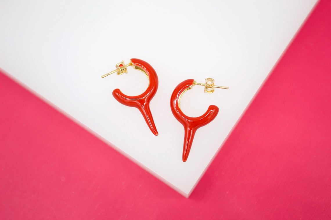 A pair of red enamel horn hoop earrings with gold-plated posts rest on a white display surface against a vibrant pink background.