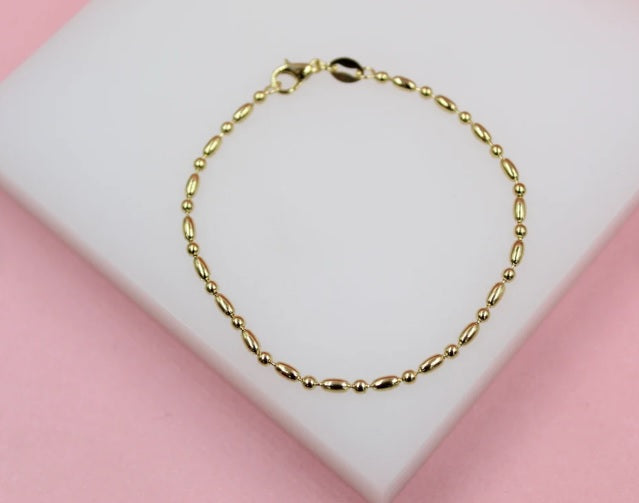 A delicate gold bead and rice-grain chain bracelet rests on a white display pad against a pink background.