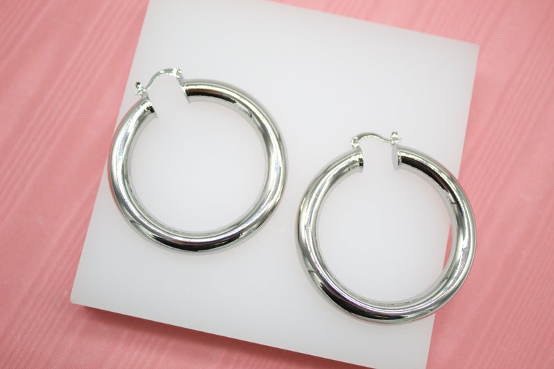 A pair of thick silver hoop earrings rests on a white display surface against a pink background.