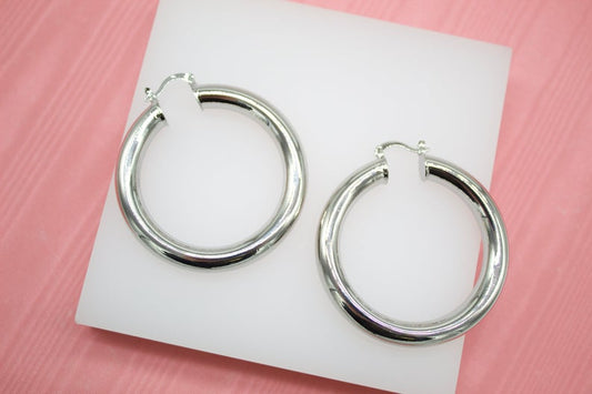 A pair of thick silver hoop earrings rests on a white display surface against a pink background.
