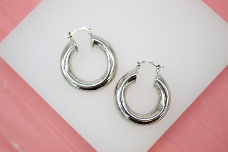 A pair of polished thick silver hoop earrings rests on a white display surface against a pink background.