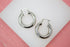 A pair of polished thick silver hoop earrings rests on a white display surface against a pink background.