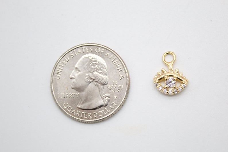 A gold evil eye charm with sparkling cubic zirconia stones rests next to a US quarter for scale.