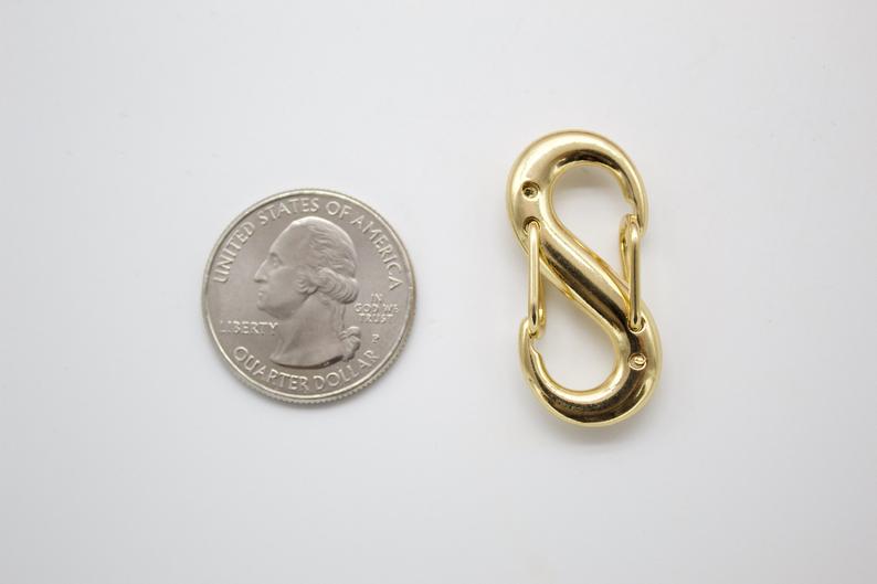 A polished gold-tone S-hook clasp rests beside a US quarter for scale on a neutral surface.