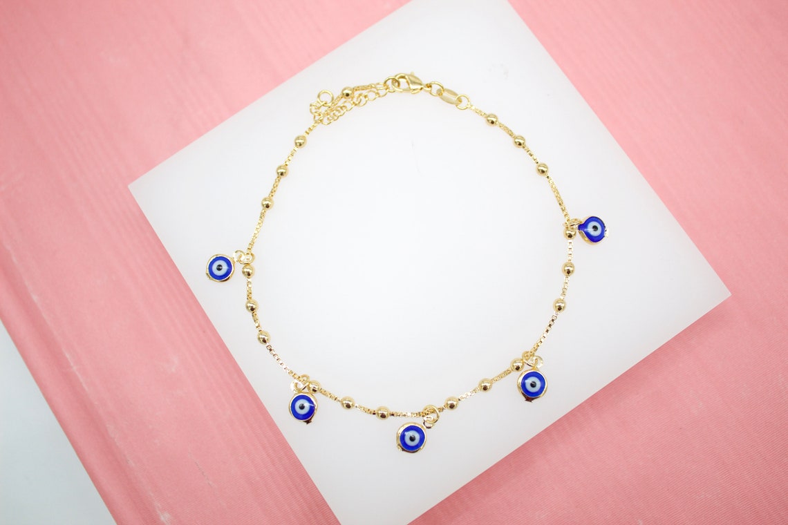 A delicate gold chain anklet featuring small evil eye charms rests on a white display surface against a pink background.