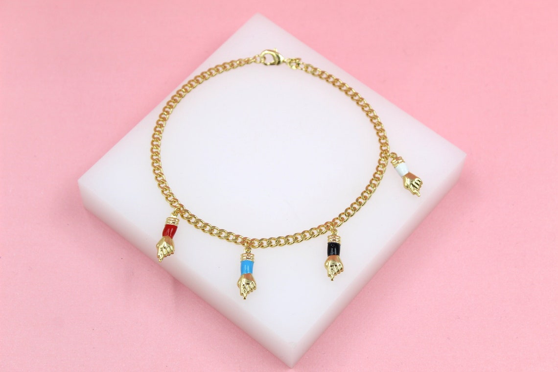 A delicate gold chain bracelet features dangling figa charms with colorful enamel details resting on a white display block against a soft pink background.