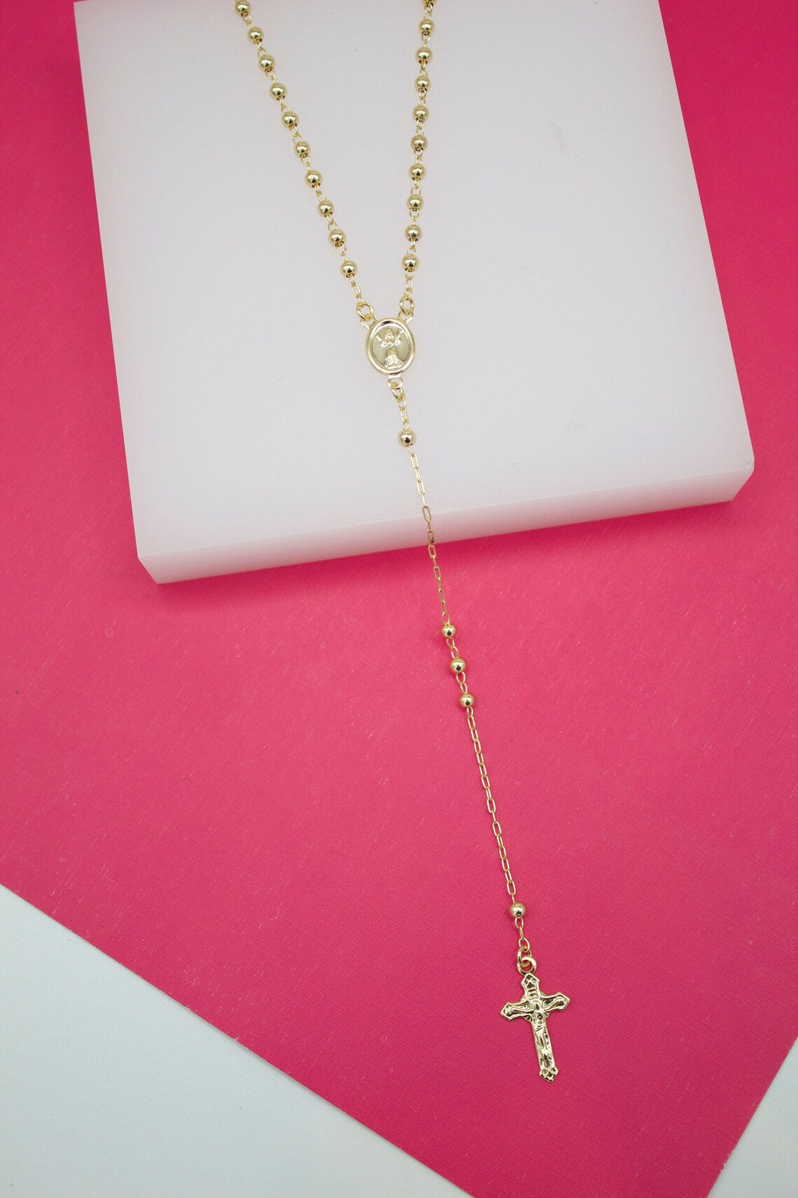 A delicate gold rosary necklace rests elegantly on a white display block against a vibrant pink background.