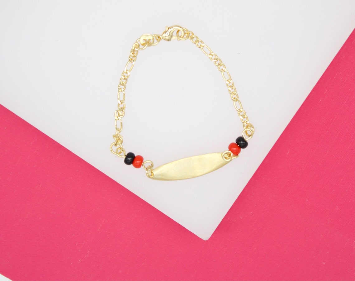 A gold ID bracelet rests on a white and pink background, featuring a blank nameplate flanked by small black and red beads.