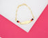 A gold ID bracelet rests on a white and pink background, featuring a blank nameplate flanked by small black and red beads.