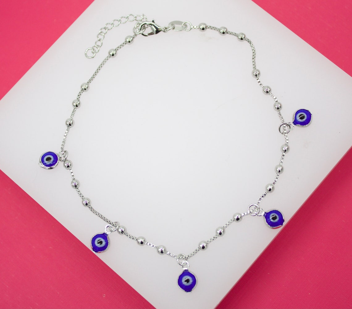 A silver evil eye anklet with blue charms rests on a white display against a vibrant pink background.