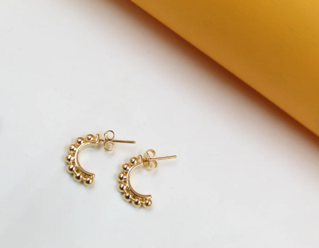 A pair of gold-plated beaded half-hoop earrings rests against a minimalist white and yellow background.