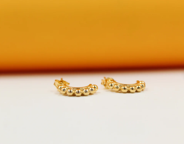 A pair of small gold huggie hoop earrings featuring a beaded texture rests against a warm orange background.