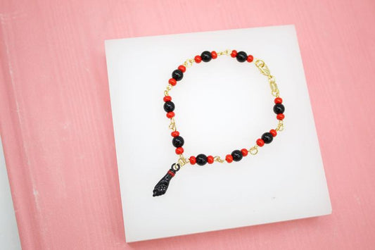 A delicate gold chain bracelet features alternating red and black beads and a black figa charm, displayed on a white surface against a pink background.