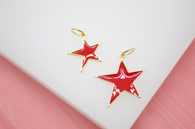 Two glossy red enamel star charms with gold trim and jump rings rest on a white display surface.
