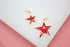 Two glossy red enamel star charms with gold trim and jump rings rest on a white display surface.