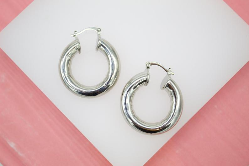 A pair of thick, polished silver hoop earrings rests on a white and pink display surface.