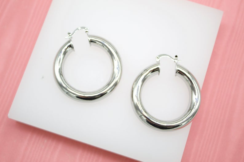 A pair of shiny silver chunky hoop earrings rests on a white display stand.