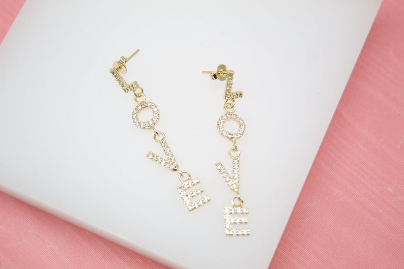 A pair of gold drop earrings spelling out the word "LOVE" with shimmering rhinestone embellishments rests on a white display surface.