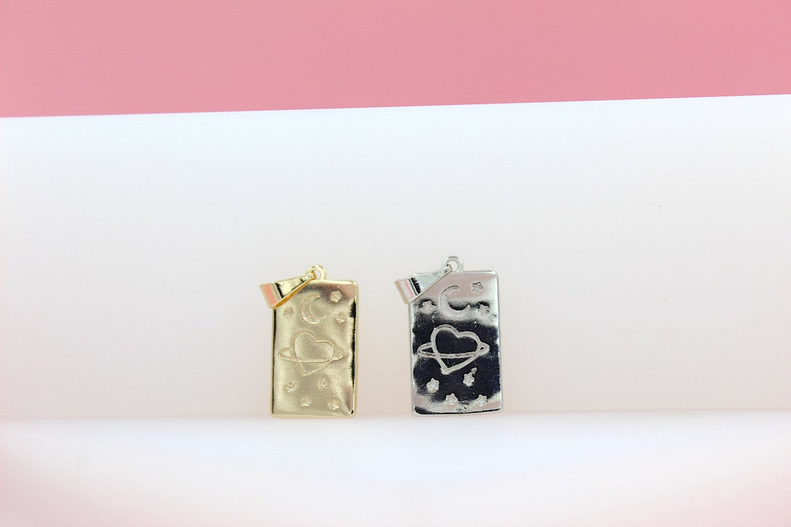 Two rectangular celestial pendants featuring engraved hearts, moons, and stars rest side-by-side on a white surface against a pink background, with the gold pendant on the left and the silver pendant on the right.