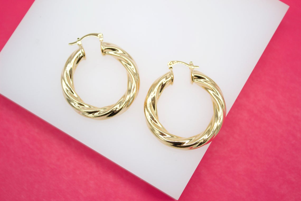 A pair of thick gold hoop earrings rests on a white display against a vibrant pink background.