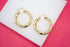 A pair of thick gold hoop earrings rests on a white display against a vibrant pink background.