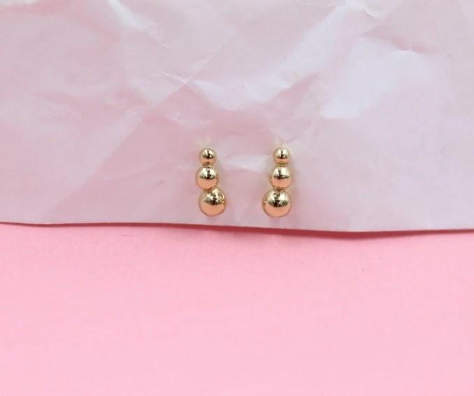 A pair of minimalist gold ball cluster stud earrings rests delicately against a textured pink and white background.
