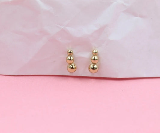A pair of minimalist gold ball cluster stud earrings rests delicately against a textured pink and white background.