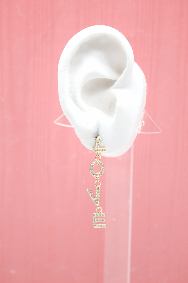 A white mannequin ear displays a dangling gold "LOVE" rhinestone letter earring against a pink background.
